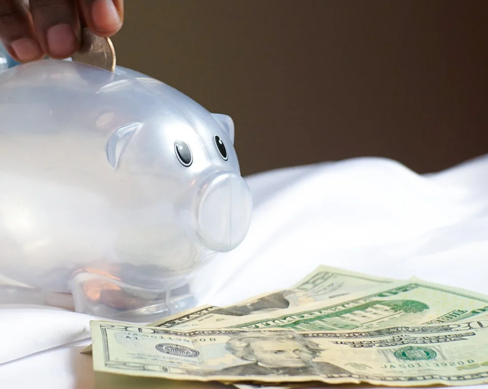 Plastic piggy bank on a desk with three 20-dollar bills in front. Hand, fingertips visible, tossing a quarter into the piggy bank.